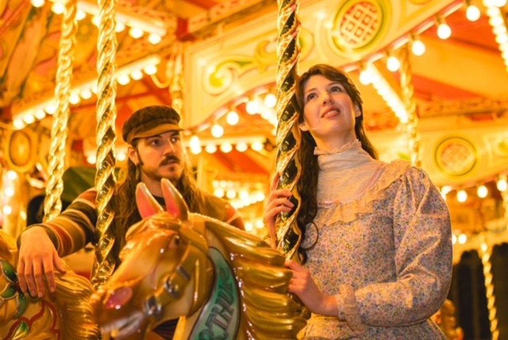 Actors in period costumes riding a carousel in a production of Rodgers and Hammerstein’s Carousel.